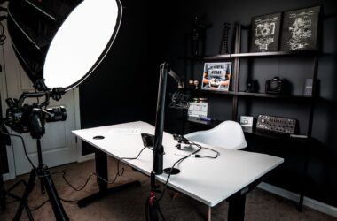 A small home vlogging studio with black walls, a large octabox softbox, a camera on a tripod, a boom-arm microphone, and a white desk