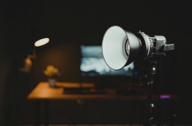 A COB studio light on a stand glowing in a dark room, with a desk, monitor, and warm lamp blurred in the background