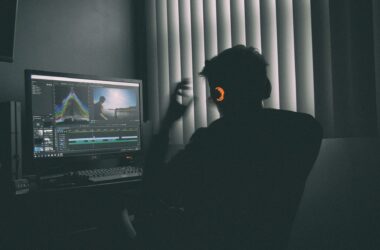 An editor silhouetted in a dark room, working at a monitor showing a video timeline and a waveform scope