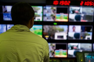 A man seen from behind watching a wall of monitors displaying video feeds and timecode in a dark control room