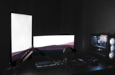 Dual glowing monitors, a backlit keyboard, and a desktop PC tower with visible internals on a black desk in a dark room