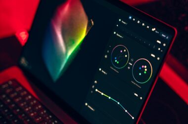 A laptop screen in a dark, red-lit room showing a three-dimensional color gamut solid beside two color wheels and a tone curve