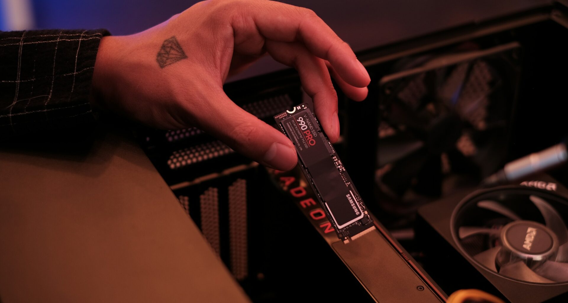 A hand holding a Samsung 990 Pro M.2 NVMe SSD above the motherboard of an open desktop PC
