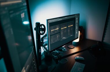 Dual monitors on a dark desk in a dimly lit room, lit by a warm lamp, with monitor arms, a boom arm and cables visible