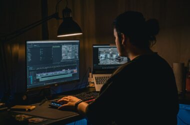 A video editor working at a dark desk lit by a single lamp, an NLE timeline on the monitor and a laptop beside it