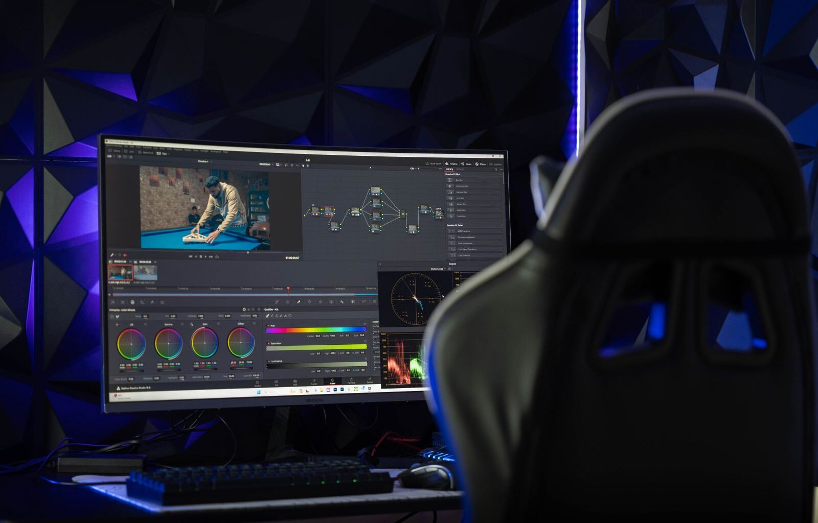 Two monitors on a dark desk showing a video editing timeline and scopes under colored mood lighting