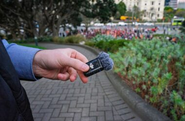 A hand holding a small wireless microphone transmitter with a furry windshield outdoors