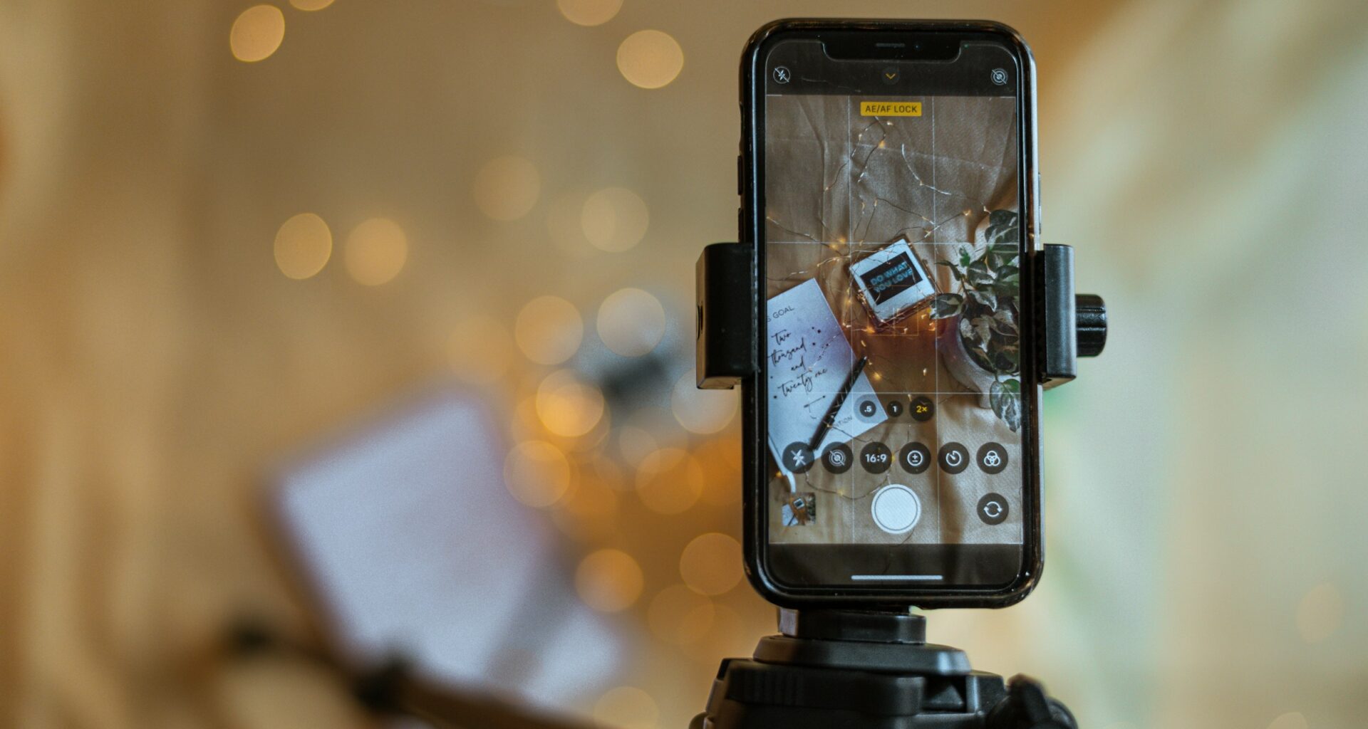 A phone mounted on a small tripod recording a flat-lay scene with warm bokeh lights behind it