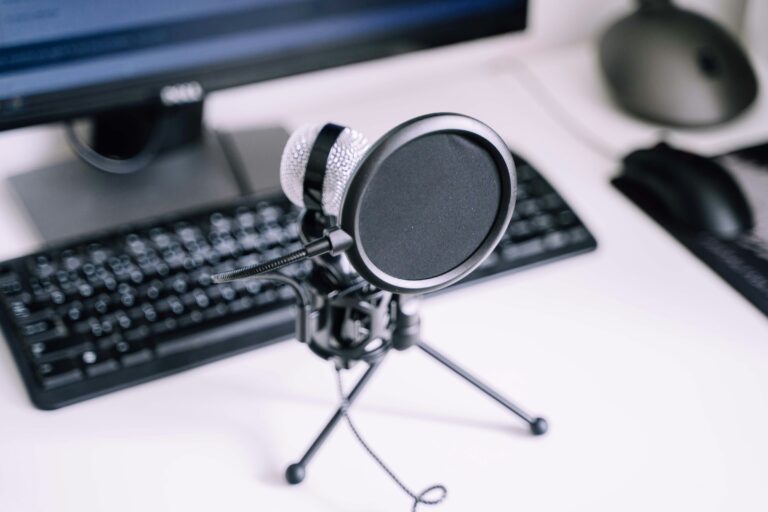 A USB microphone with a pop filter on a desk in front of a keyboard and monitor