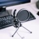 A USB microphone with a pop filter on a desk in front of a keyboard and monitor