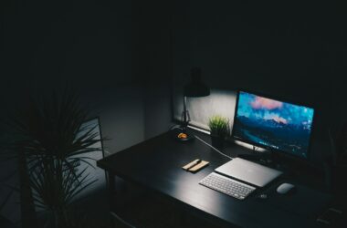 A dark desk with a monitor, laptop, keyboard, and desk lamp