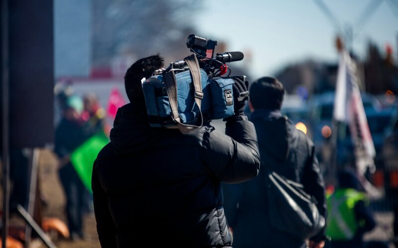 A news camera operator with a shoulder-mounted ENG camera filming at an outdoor event
