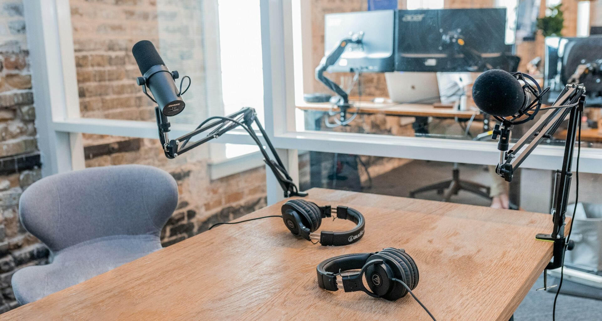 Two broadcast microphones on boom arms and two pairs of headphones on a wooden podcast table
