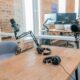 Two broadcast microphones on boom arms and two pairs of headphones on a wooden podcast table